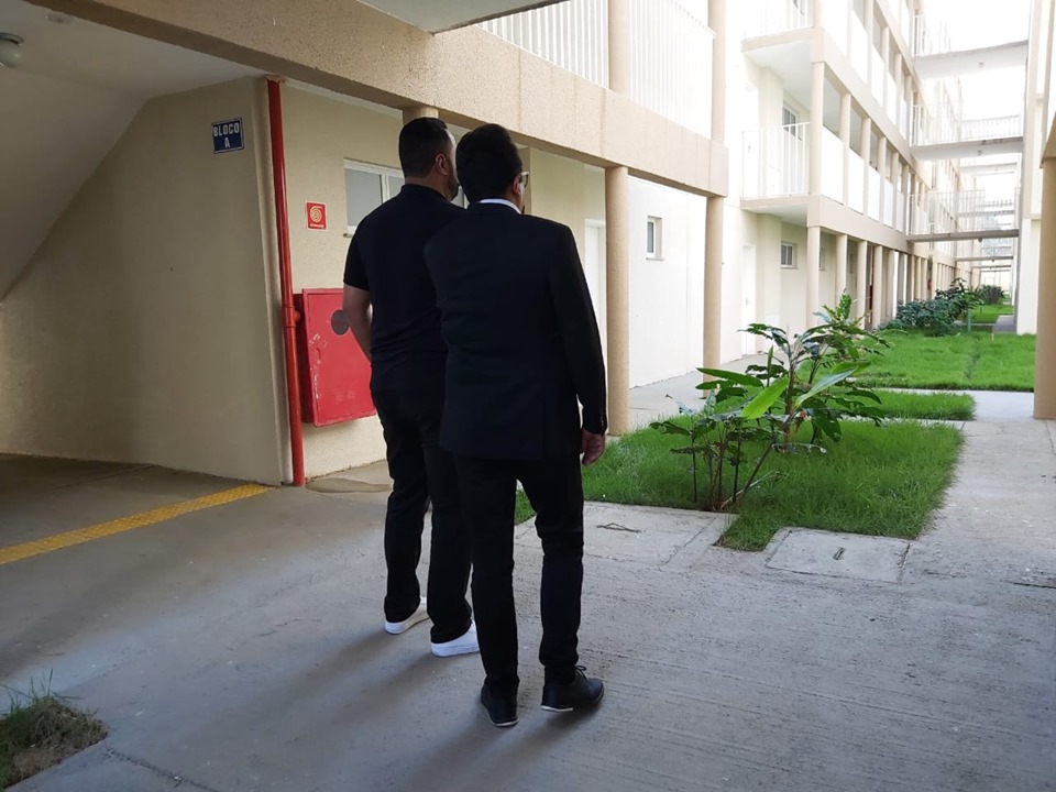 Dois homens, vestidos com calças e blusas pretas, observam a área externa do Conjunto habitacional Academia Paulista.